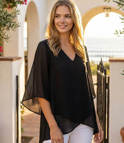 Woman wearing a black top and white pants standing outdoors with a scenic background.