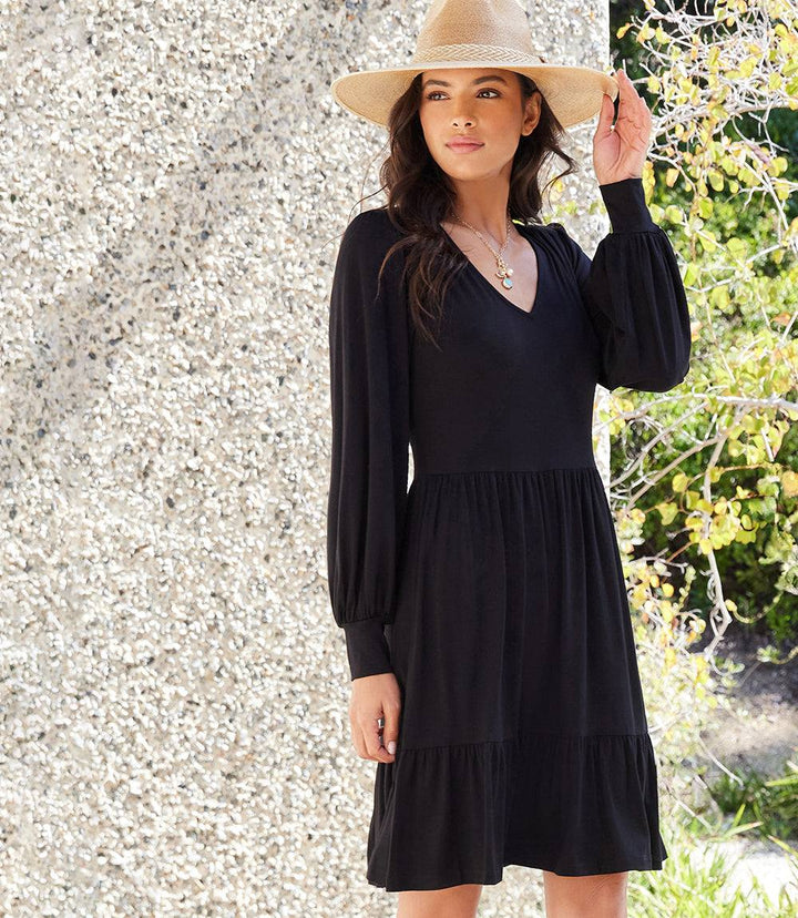 Woman wearing a black long sleeve tiered dress with a V-neckline and gathered cuffs, styled with a beige hat outdoors.