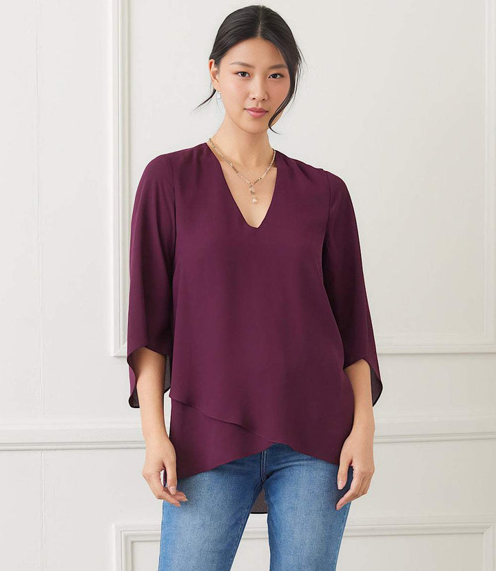 Woman wearing a deep purple Bracelet Sleeve Asymmetric Top with V-neck, flare sleeves, and layered asymmetrical hem paired with blue jeans.