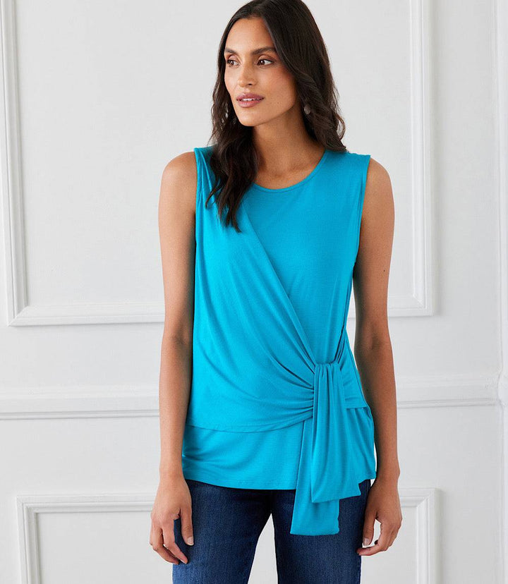 Woman wearing a sleeveless turquoise side drape top with a scoop neck and side tie detail paired with dark jeans in a white room