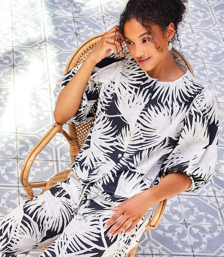 Woman wearing black and white palm print puff sleeve button-front shirt sitting on a wicker chair