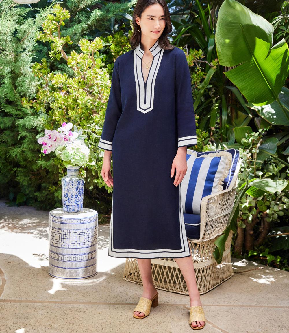 Woman wearing The St. Tropez Midi Dress in navy linen blend with white ribbon edges, standing outdoors near wicker chair and blue patterned ceramic stool