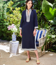 Woman wearing navy St. Tropez Midi Dress with white ribbon edges, standing outdoors near a wicker chair and plants