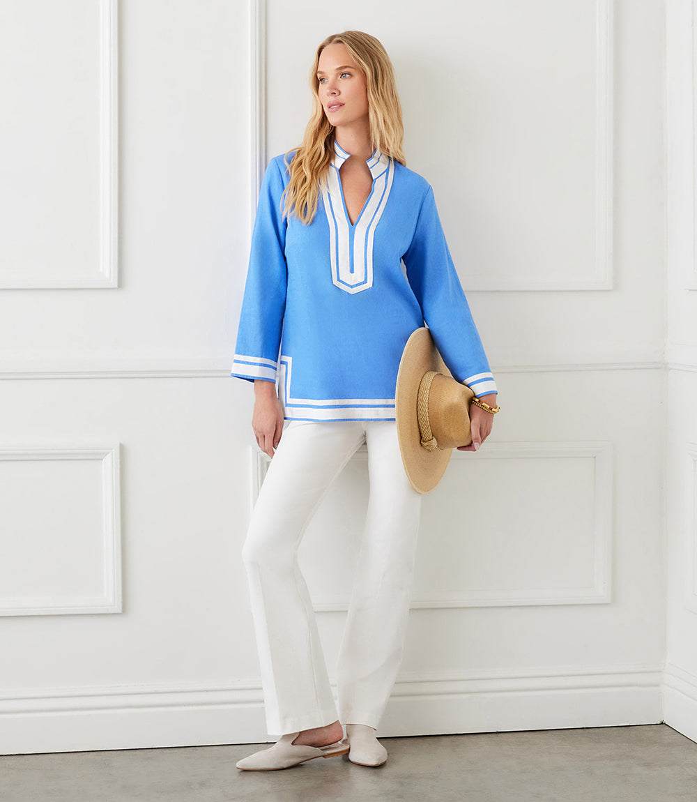 Woman wearing blue The St. Tropez Tunic with white grosgrain ribbon trim, paired with white pants and holding a hat indoors