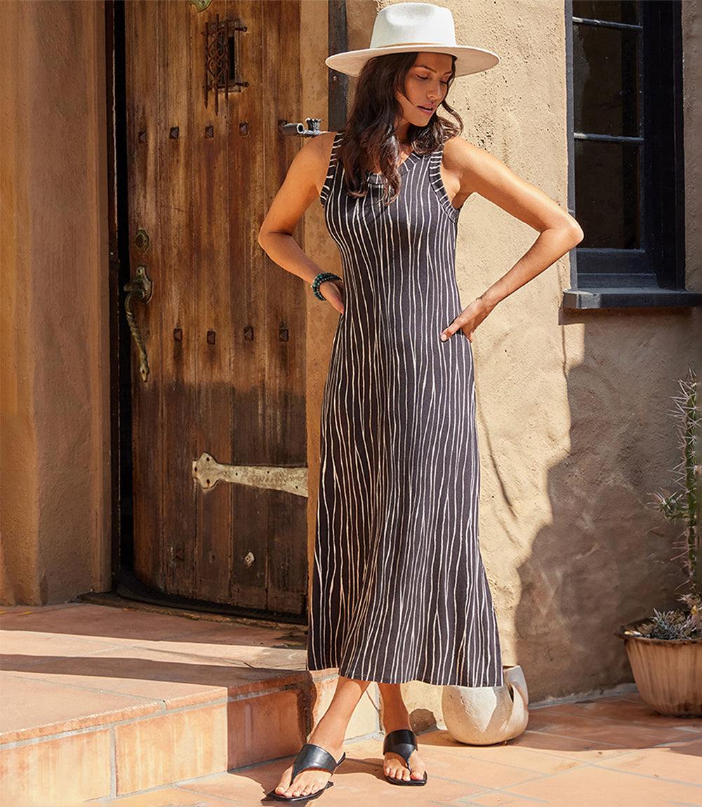 Woman wearing sleeveless midi dress with wavy vertical stripes and scoop neck, paired with hat and sandals.