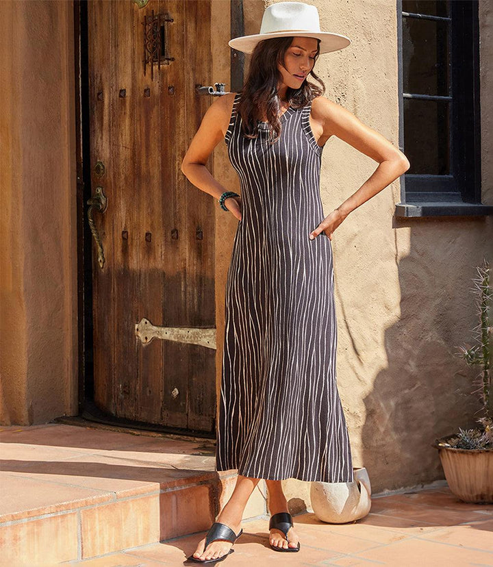 Woman wearing sleeveless midi dress with wavy vertical stripes and scoop neck, paired with hat and sandals.