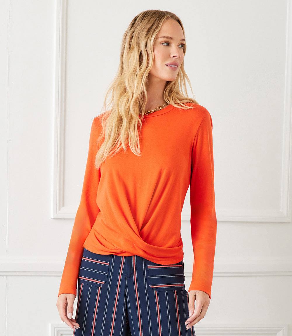 Woman wearing orange long sleeve drape front top with twist hem and crew neck paired with striped pants.