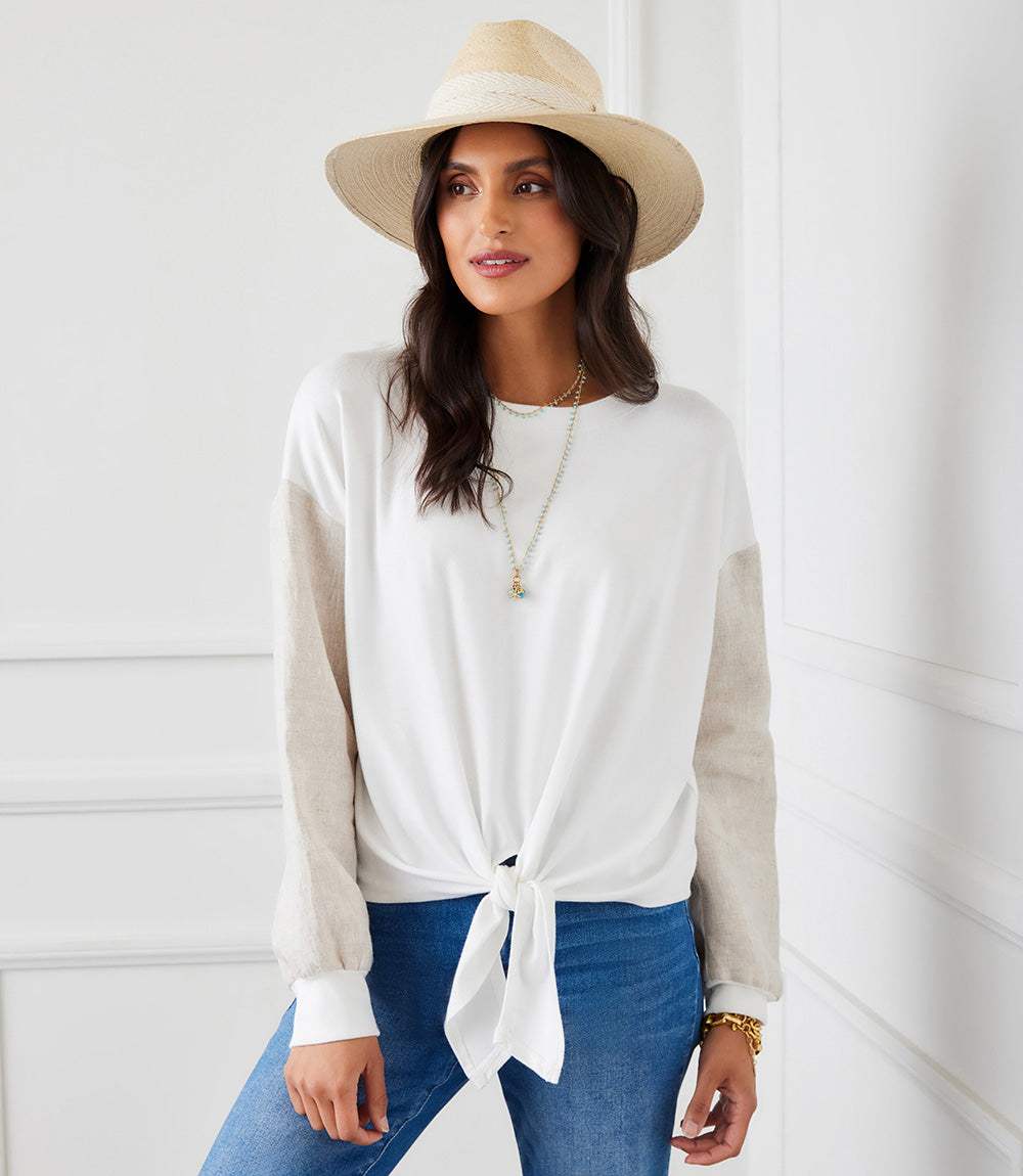 Woman wearing a sustainable contrast tie-front top with white body and beige linen sleeves, styled with blue jeans and a hat.