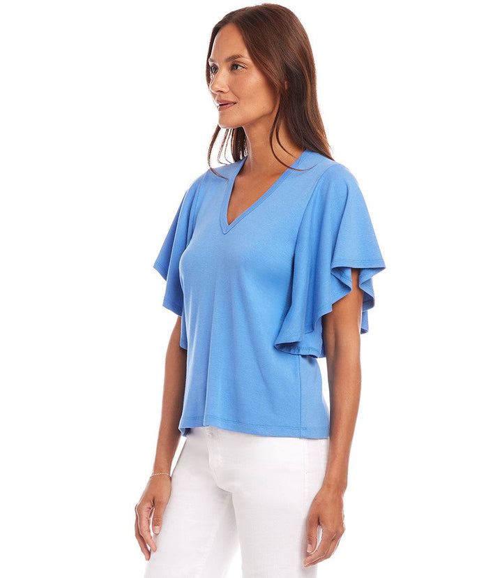 Woman wearing a blue flutter sleeve V-neck top made from organic cotton paired with white pants