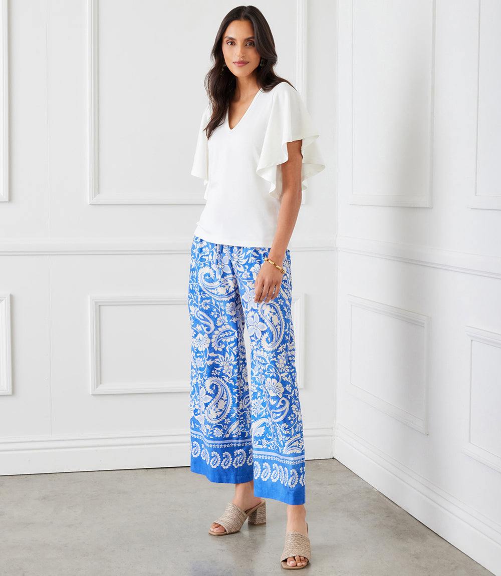 Woman wearing white flutter sleeve v-neck top paired with blue patterned wide-leg pants in bright room