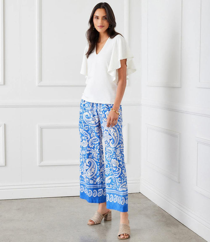Woman wearing white flutter sleeve v-neck top paired with blue patterned wide-leg pants in bright room