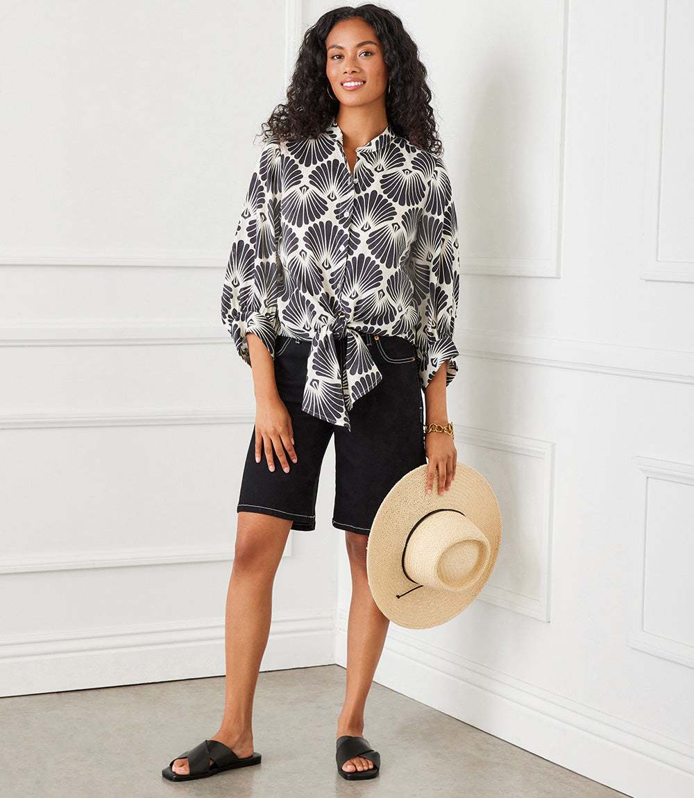 Woman wearing a black and cream blouson sleeve linen tie-front top with sea shell print, paired with black shorts and holding a straw hat.