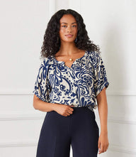 Woman wearing a short sleeve peasant top with navy paisley print and puff sleeves paired with dark pants