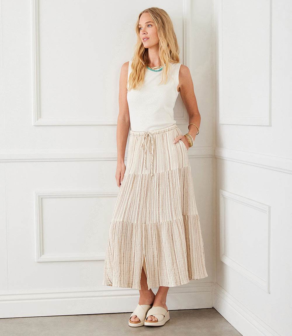 Woman wearing a metallic stripe tiered midi skirt with cream and tan stripes and gold metallic thread, paired with a sleeveless white top and sandals in a white room.