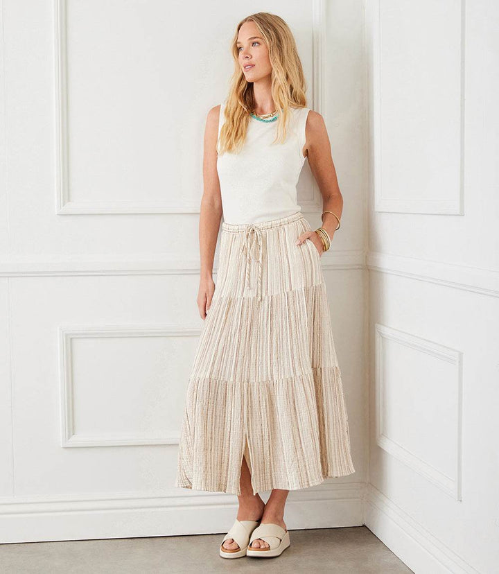 Woman wearing a metallic stripe tiered midi skirt with cream and tan stripes and gold metallic thread, paired with a sleeveless white top and sandals in a white room.