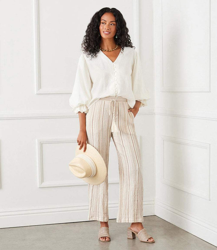 Woman wearing metallic stripe woven gauze drawstring ankle pants with a white blouse and holding a beige hat in a bright room