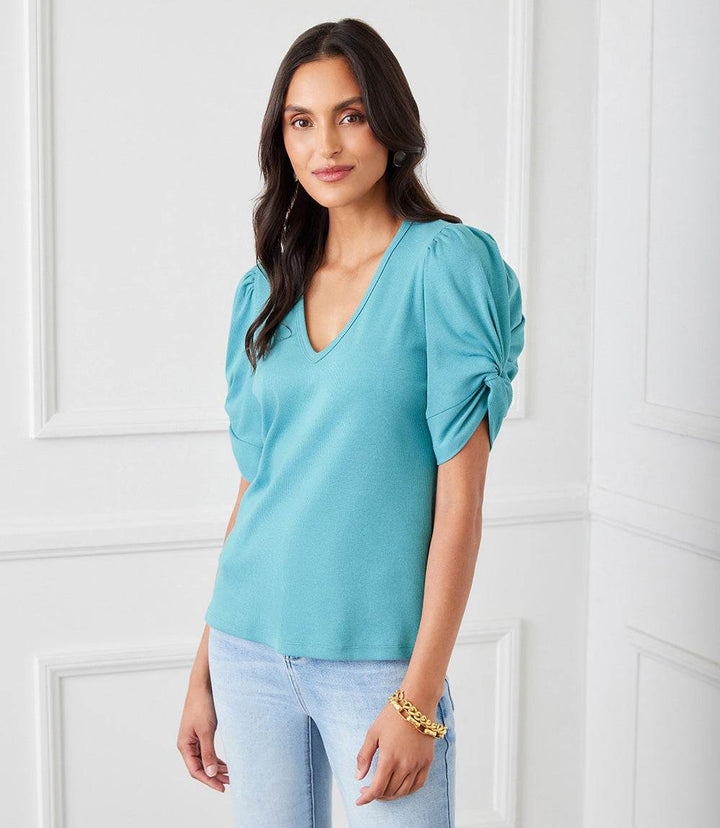 Woman wearing a teal ribbed twist sleeve V-neck top with puff sleeves and twist cuff detail paired with light blue jeans