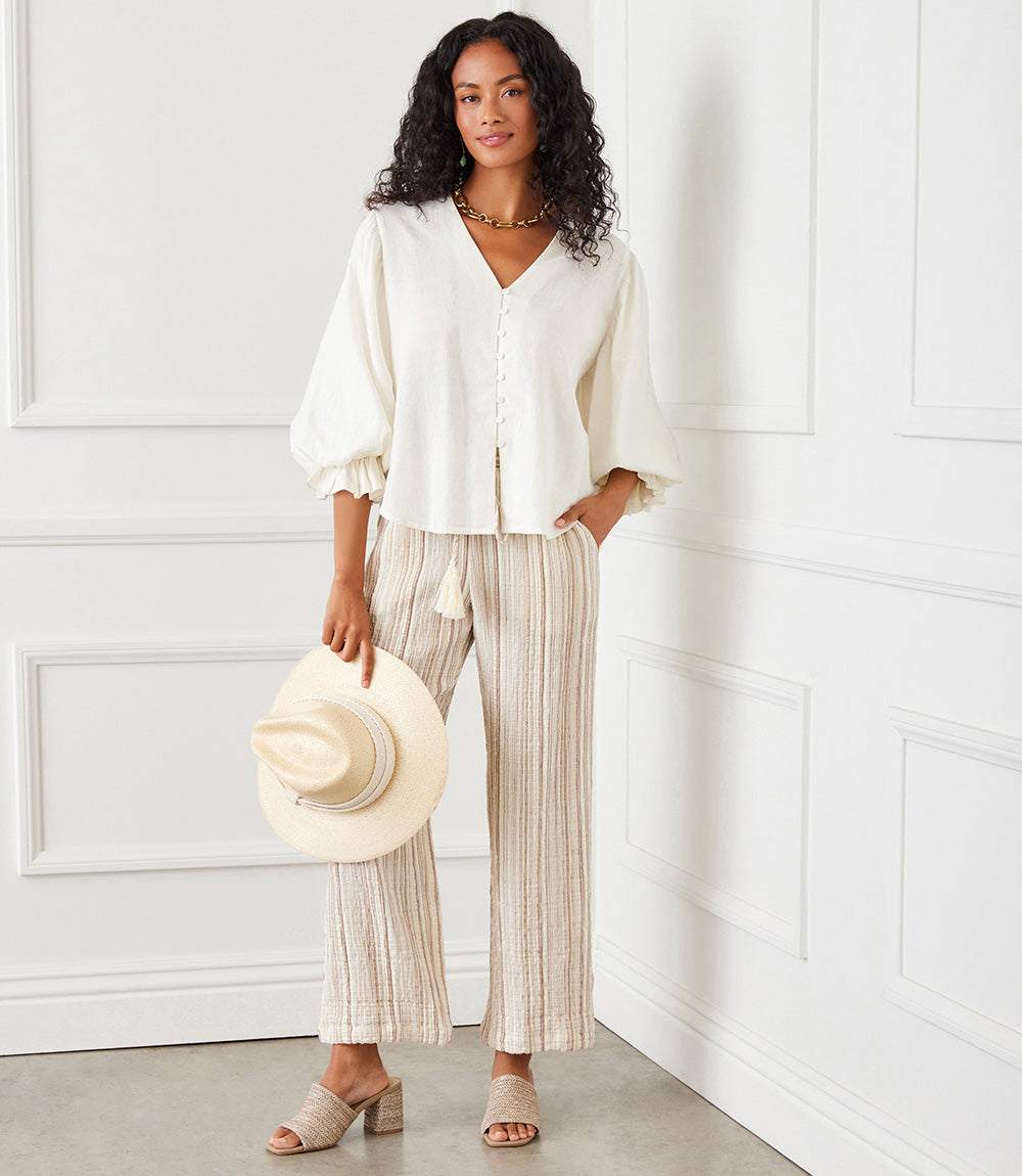 Woman wearing a white layered sleeve shirred blouse with 3/4 elasticized ruffled sleeves, beige striped wide-leg pants, holding a beige hat.