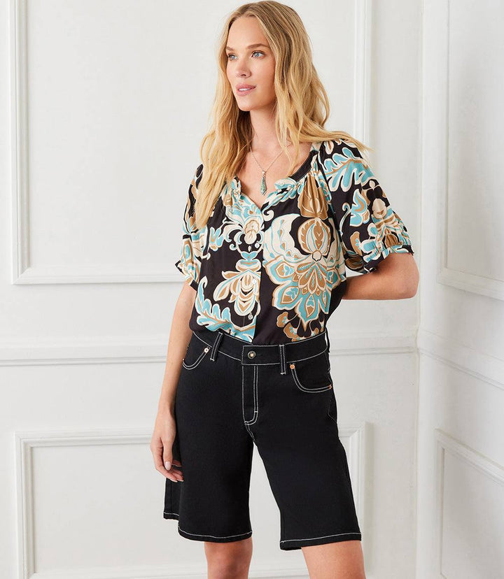 Woman wearing black denim Bermuda shorts with contrast white stitching and a floral patterned blouse standing indoors
