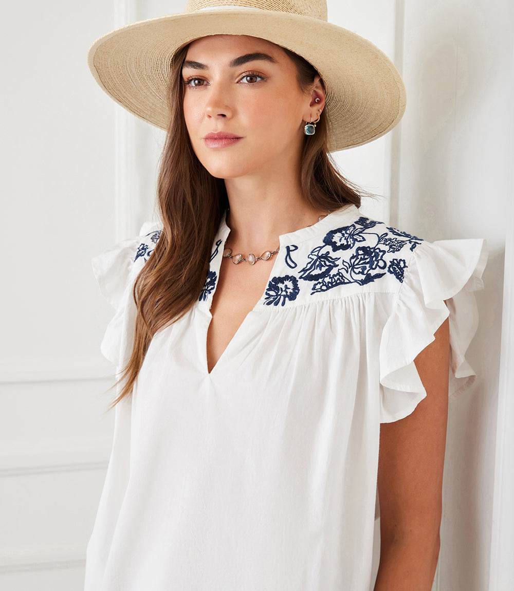Woman wearing white cotton top with navy embroidered ruffle sleeves and split neck design, accessorized with a straw hat and earrings.