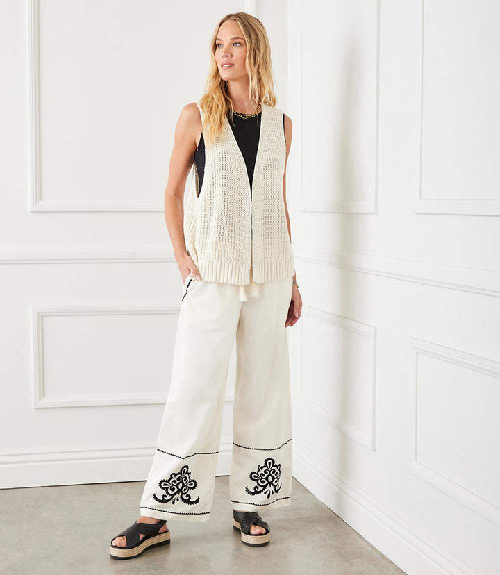 White embroidered drawstring ankle pants with wide legs and black detailing, styled with a sleeveless cream knit vest and black sandals.