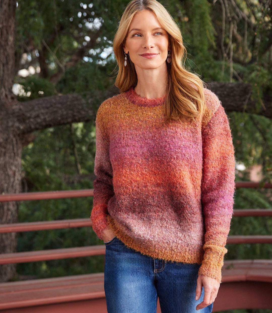 Woman wearing a loose fit ombre boucle sweater in warm shades of orange, pink, and purple with a crew neck and long sleeves.