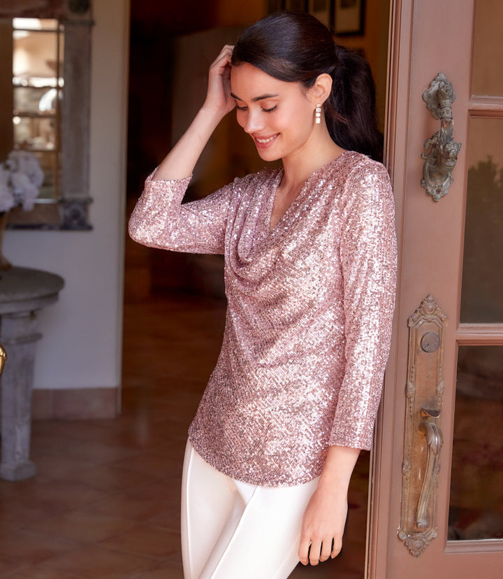 Woman wearing pink 3/4 sleeve drape front sequin top with white pants indoors