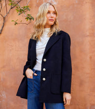 Woman wearing a black oversized brushed blazer with floral buttons and front patch pockets against a textured brown wall