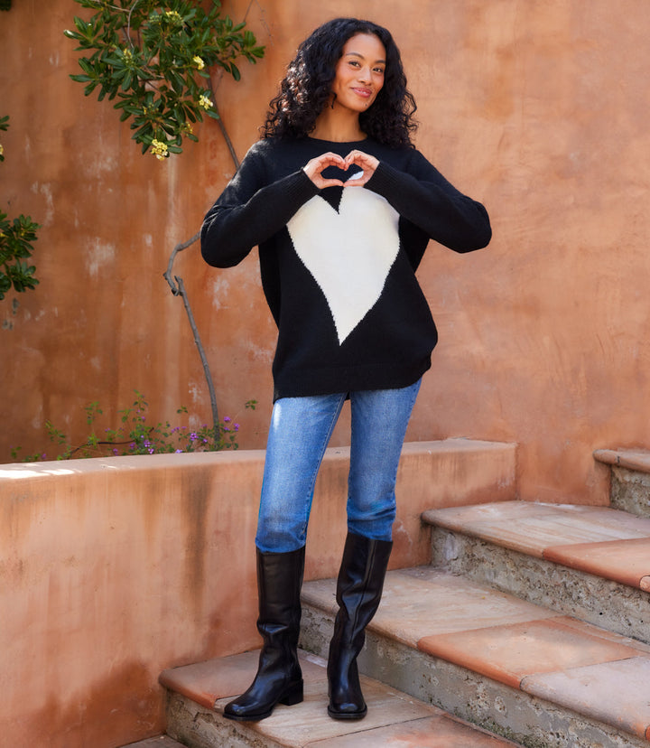 Woman wearing black Heart Jacquard Sweater with oversized white heart motif, blue jeans, and black boots outdoors