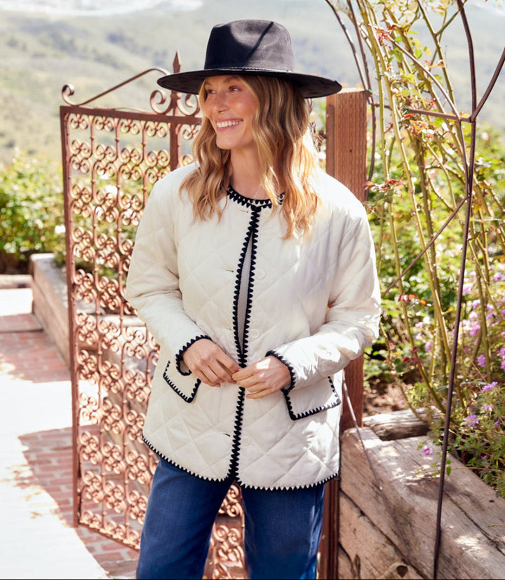 Woman wearing cream Edge Stitch Quilted Jacket with black trim, black hat, and blue jeans outdoors