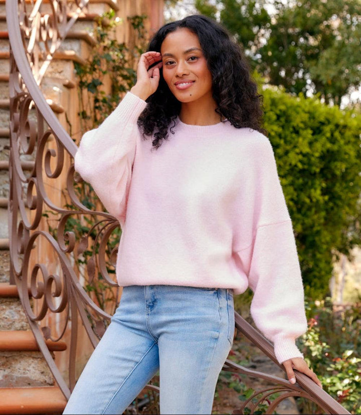 Woman wearing light pink dolman sleeve sweater with crew neck and drop shoulder design paired with light blue jeans outdoors by staircase