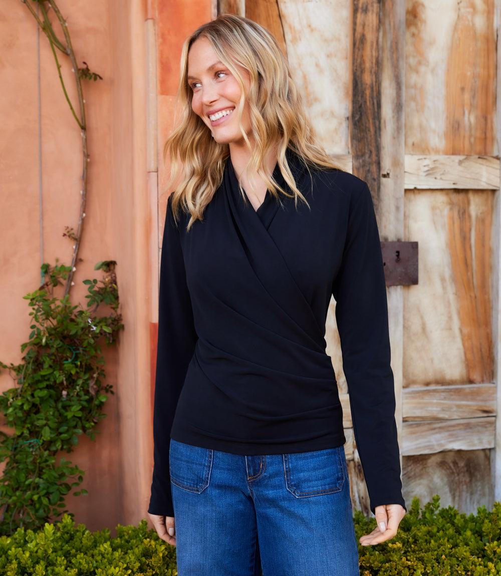 Woman wearing black travel jersey drape front crossover top with long sleeves and side pleats standing outdoors