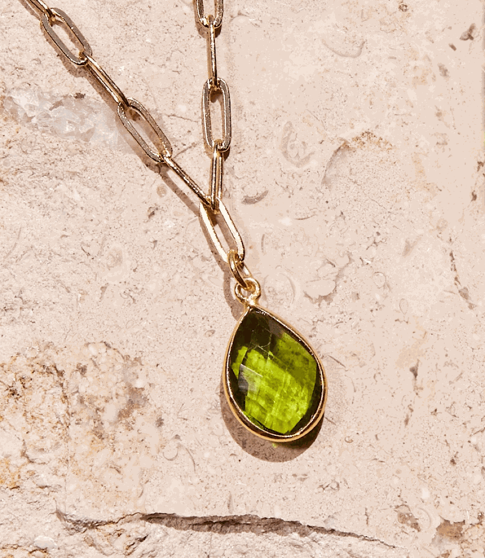 Gold plated chain necklace with a vibrant green teardrop-shaped peridot gemstone pendant on a textured stone background