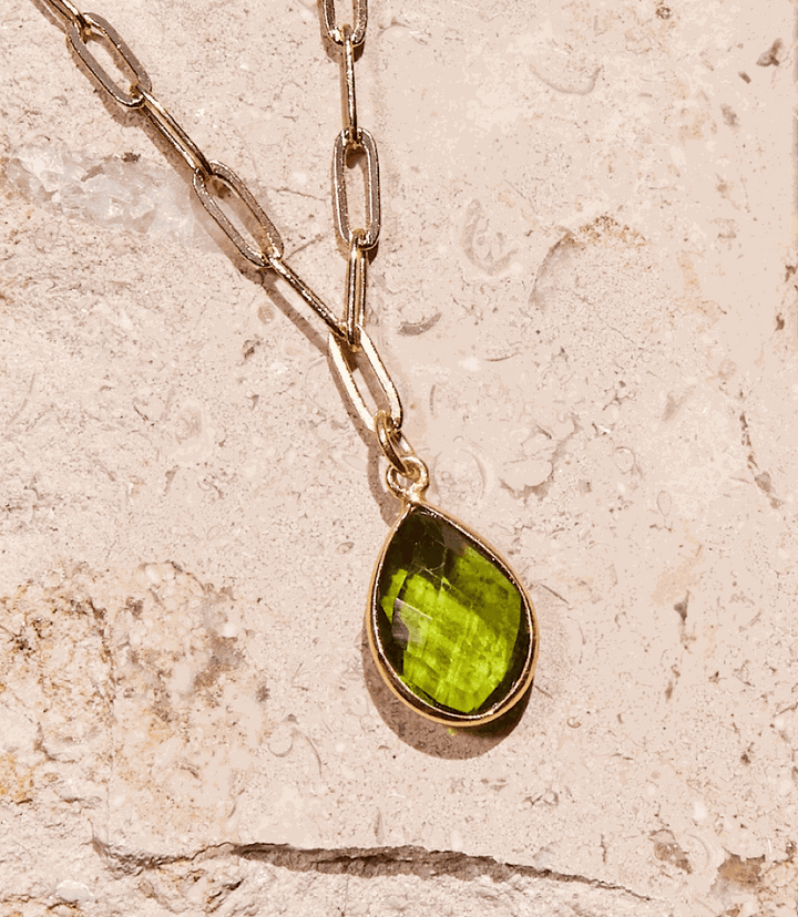 Gold plated chain necklace with a vibrant green teardrop-shaped peridot gemstone pendant on a textured stone background