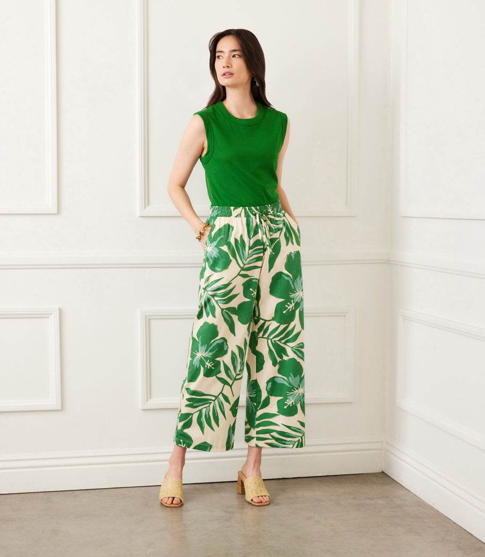 Woman wearing green floral print drawstring ankle pants with wide legs and elastic waistband paired with green sleeveless top and beige sandals in a white room