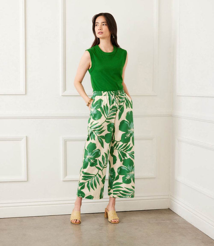Woman wearing green floral print drawstring ankle pants with wide legs and elastic waistband paired with green sleeveless top and beige sandals in a white room
