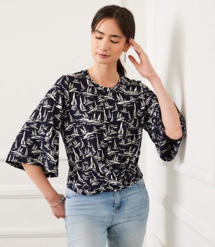 Woman wearing navy flare sleeve drape front top with cream sailboat print and light blue jeans