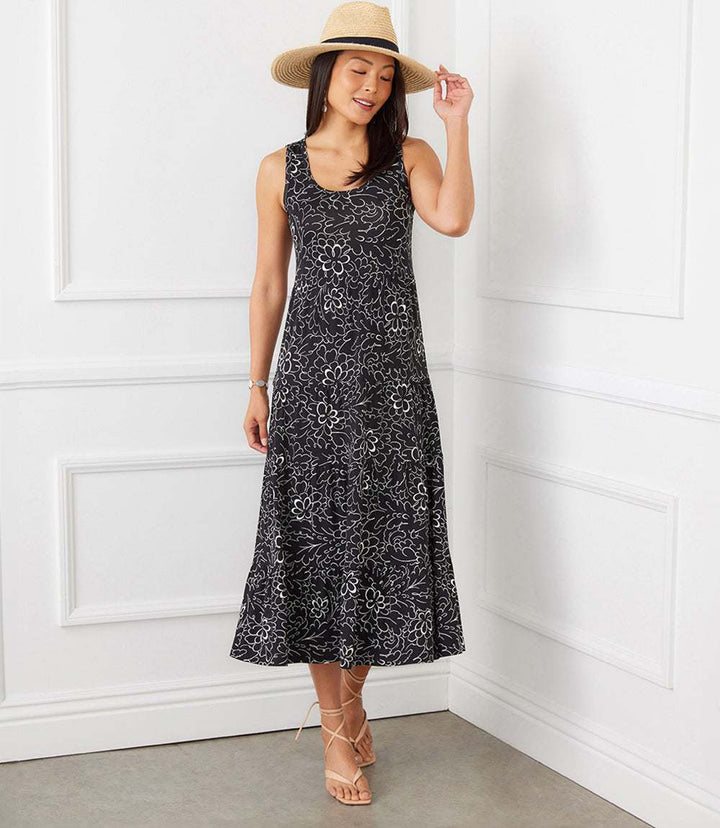 Woman wearing sleeveless black tiered midi dress with white flower print and scoop neck, accessorized with straw hat and sandals.