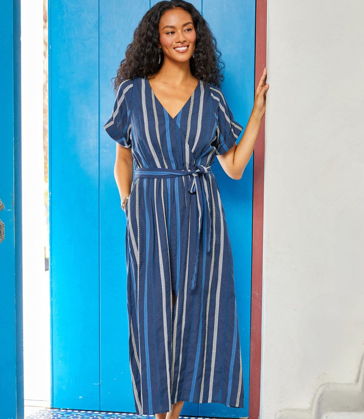 Woman wearing navy blue embroidered cuffed sleeve midi dress with vertical cream stripes and a removable waist tie belt standing against a blue door.