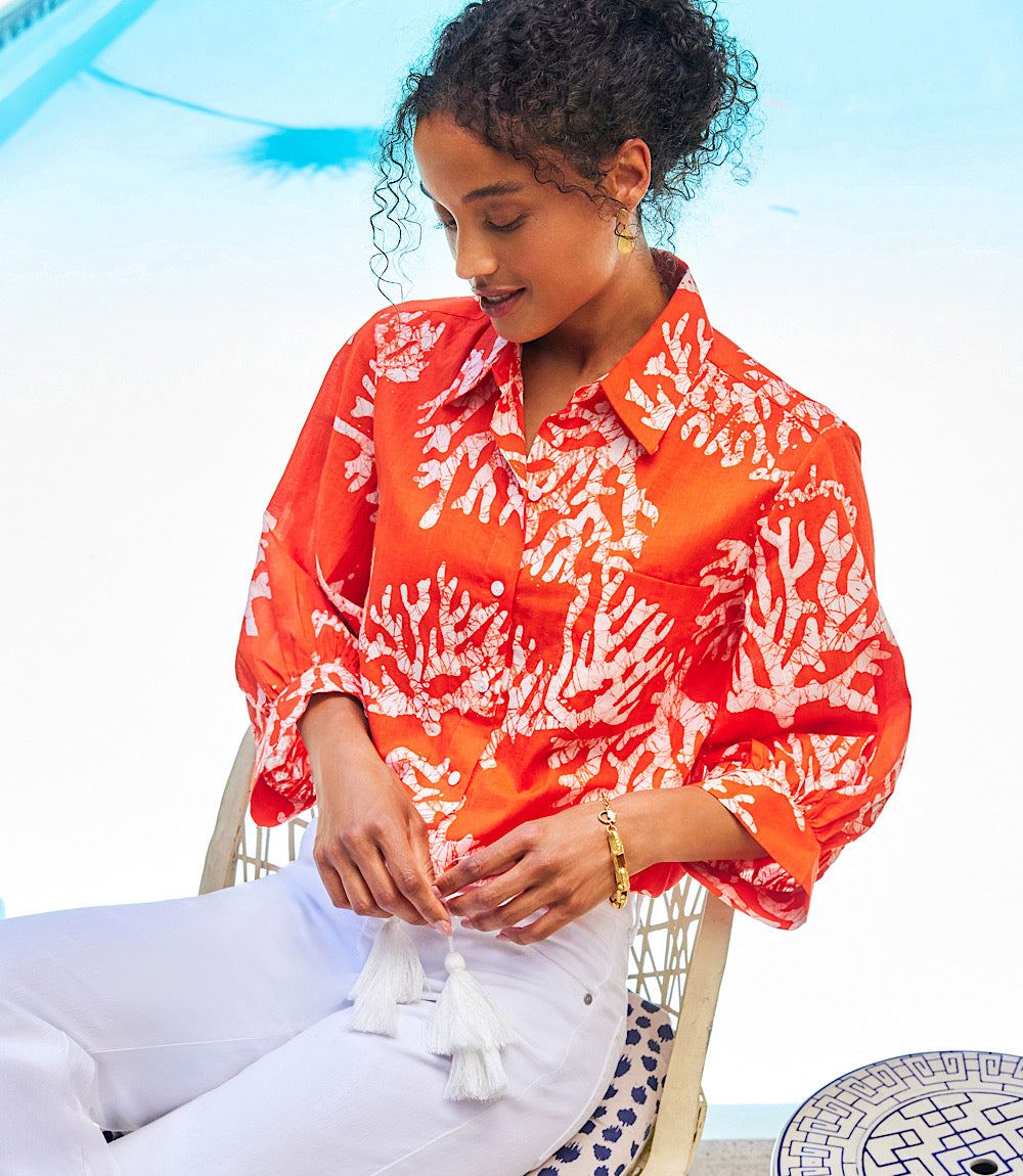 Woman wearing coral reef print tassel tie waist top with blouson sleeves and white pants sitting outdoors near pool
