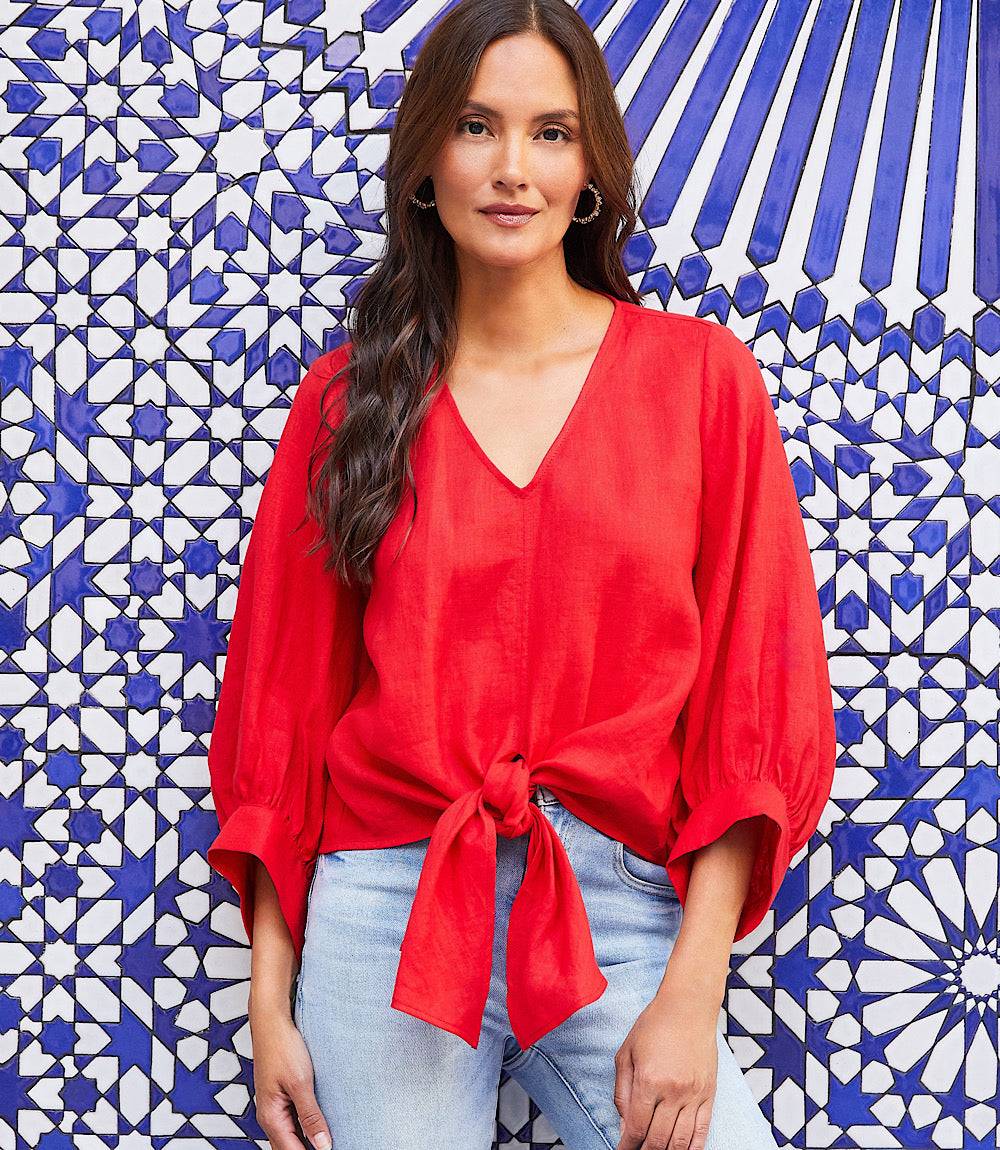 Woman wearing a red blouson sleeve linen tie-front top with V-neck and knotted front hem against blue and white patterned background
