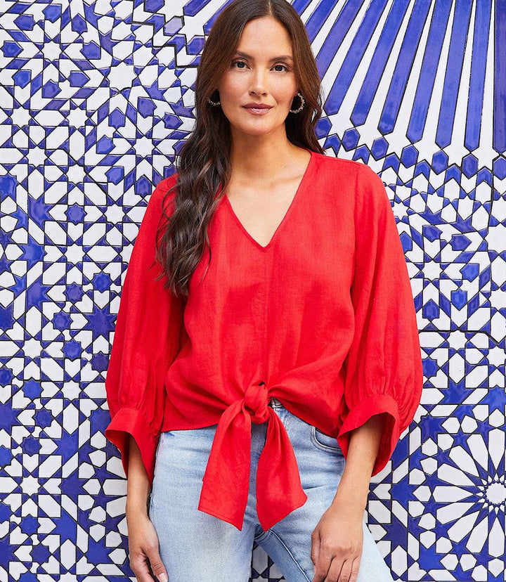 Woman wearing a red blouson sleeve linen tie-front top with V-neck and knotted front hem against blue and white patterned background