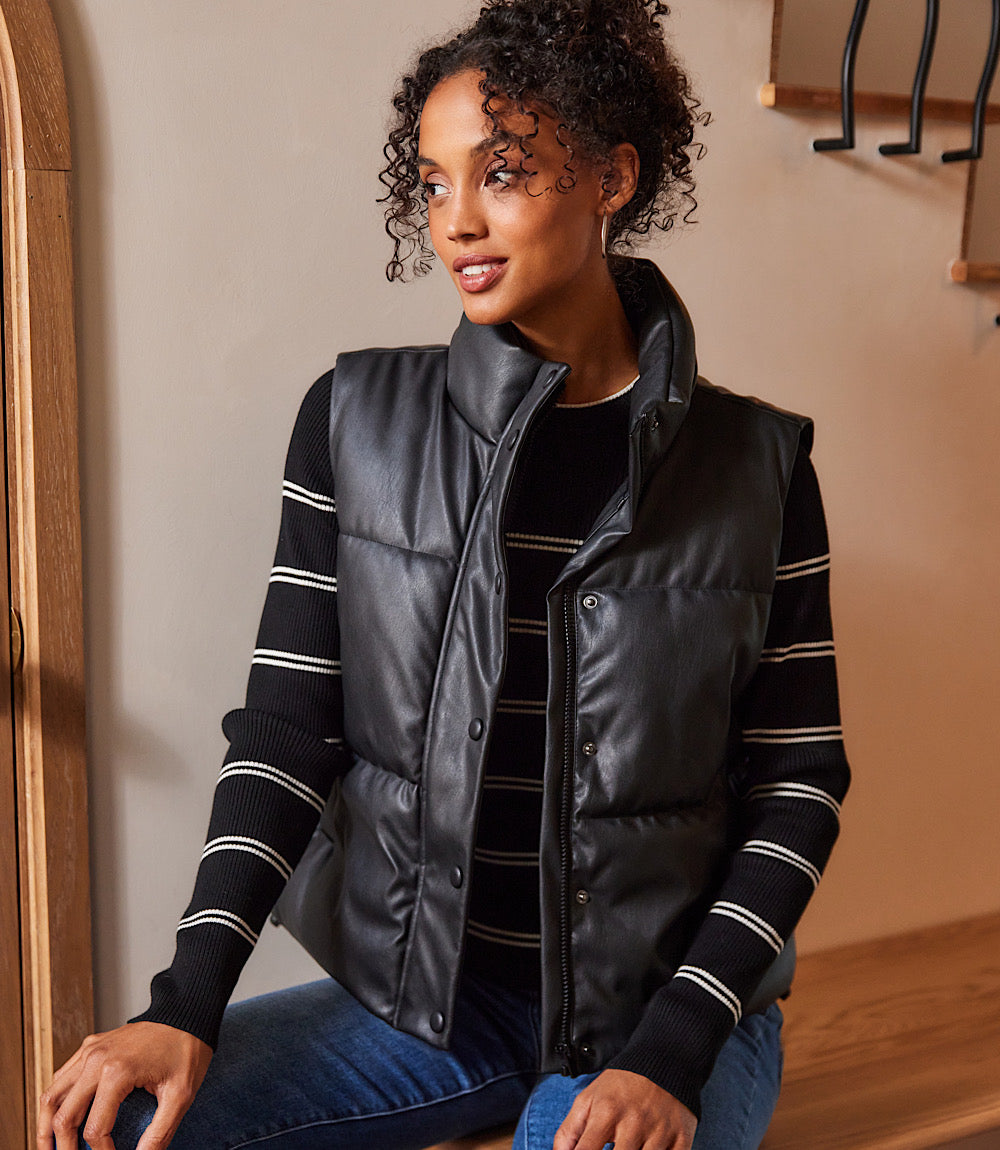 Woman wearing a black faux leather quilted puffer vest with snap and zipper closure and stand collar over a striped black sweater.