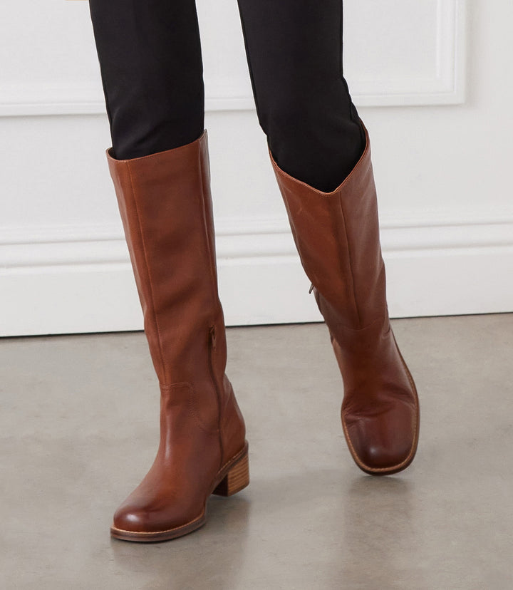 Brown leather round toe knee high boots with 2-inch heel and half zipper closure worn with black pants