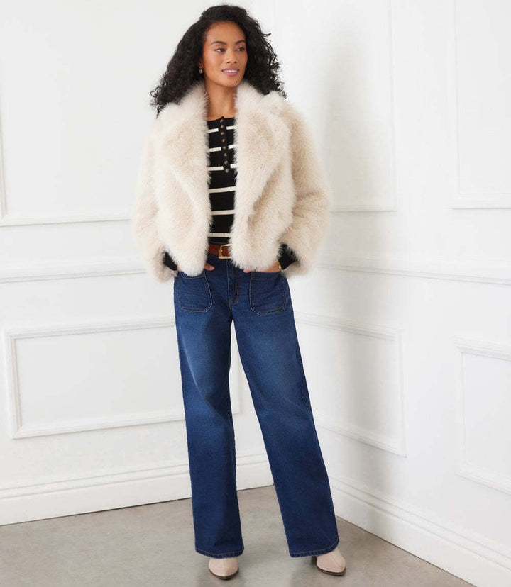 Woman wearing a cream cropped faux fur jacket with wide collar and button front paired with striped top and blue jeans