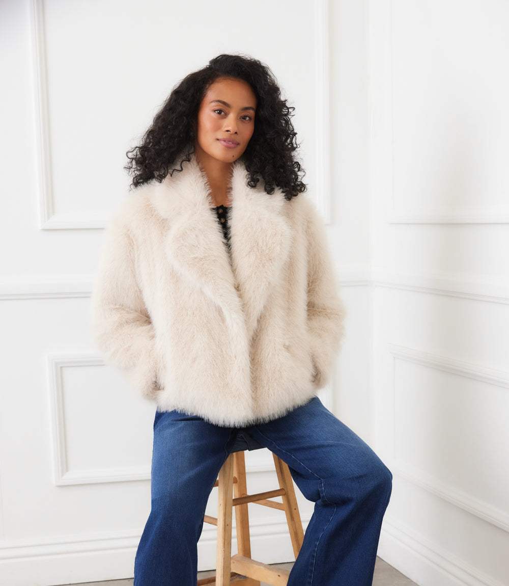 Woman wearing a cream cropped faux fur jacket with wide collar and button front closure sitting on a wooden stool