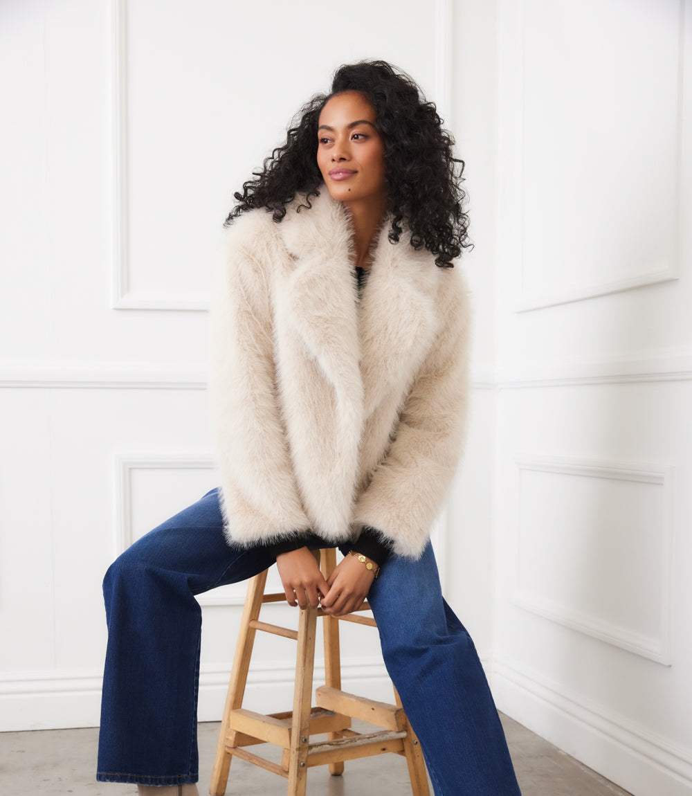 Woman wearing a cropped cream faux fur jacket with wide notched collar and button front sitting on a wooden stool