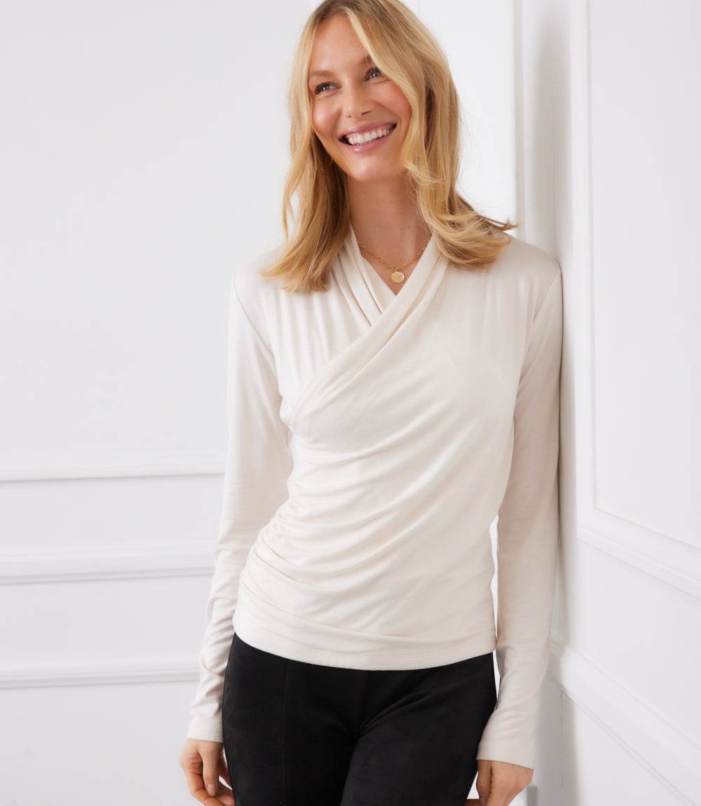 Woman wearing a cream long sleeve crossover V-neck top with side shirring and slim fit.