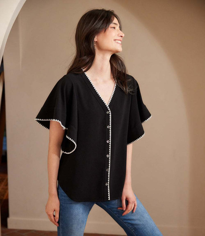 Woman wearing black flutter sleeve V-neck button-down top with cream edge stitch and shirttail hem