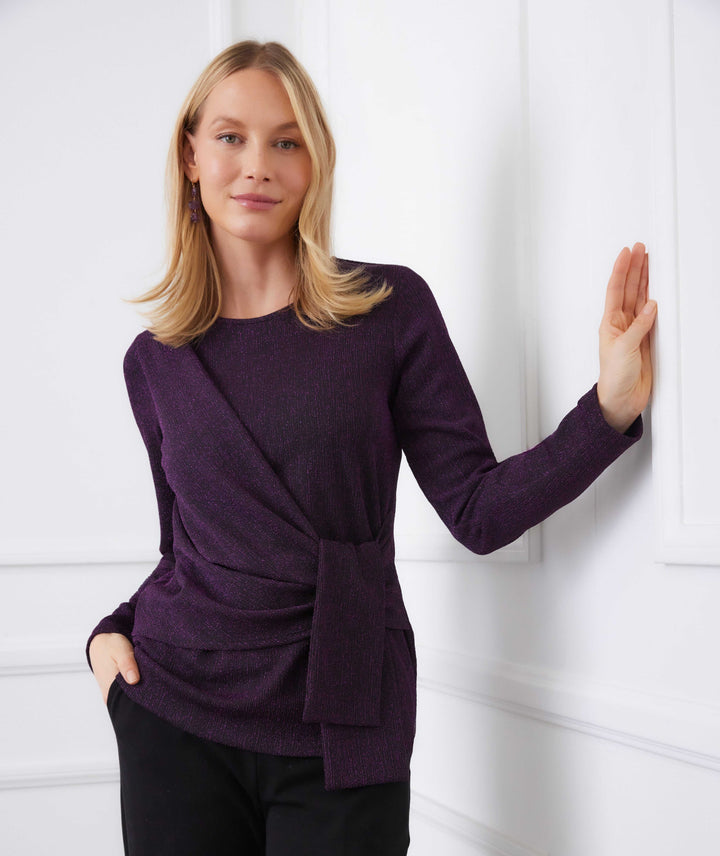Woman wearing a long sleeve metallic knit side-tie top in purple with a scoop neck and draped waist detail